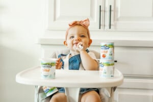 Baby girl sitting in her high chair eating plain Stonyfield yogurt with cups of yogurt around her on the tray.