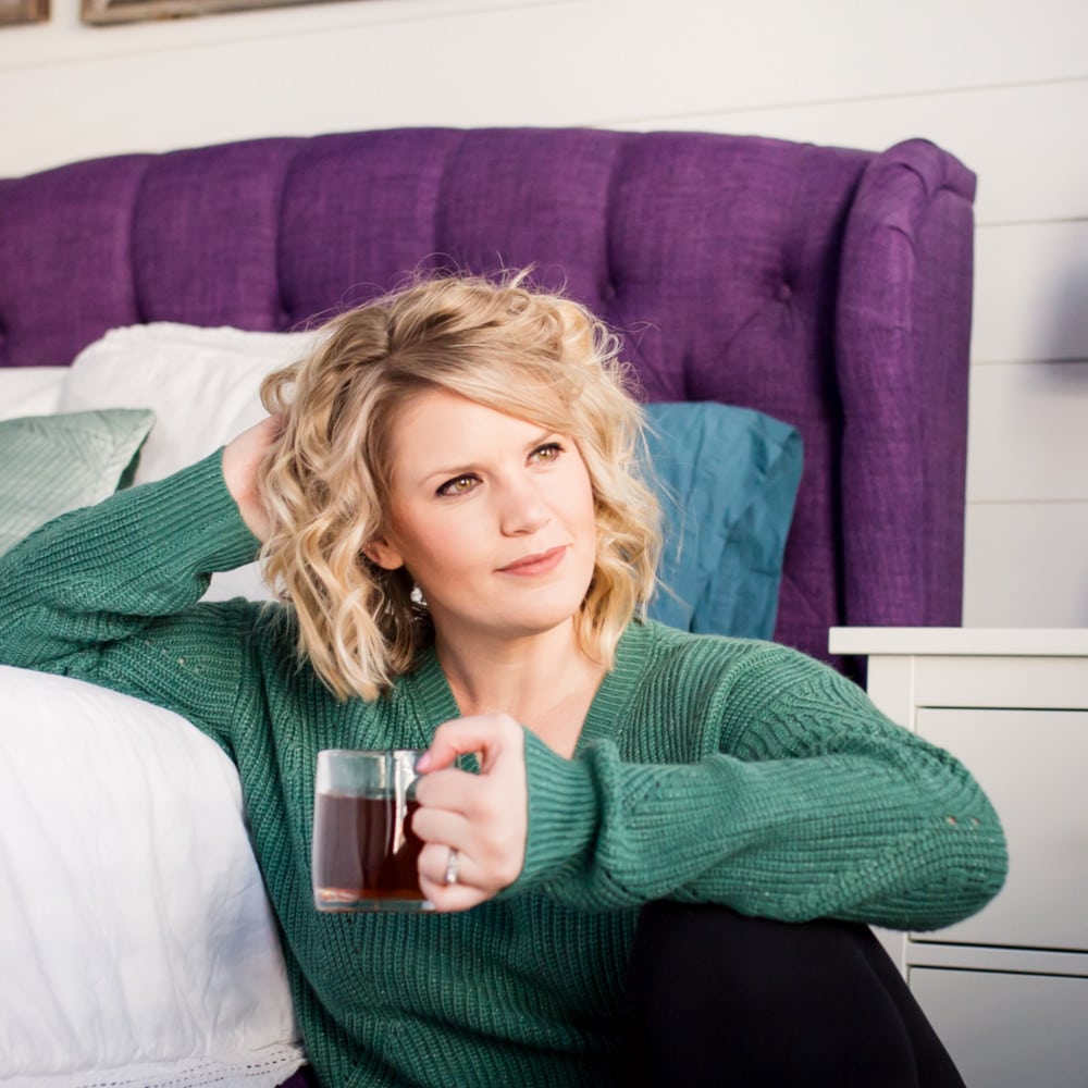 Meghan Quinn sitting on the floor next to a bed holding a cup of coffee.