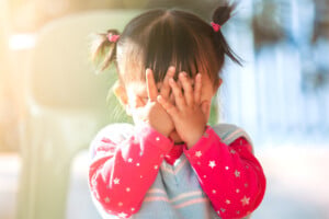 A shy child with two small pigtails, wearing a pink star-patterned sweater and light blue vest, covers their face with their hands. The background is blurred with sunlight filtering through.