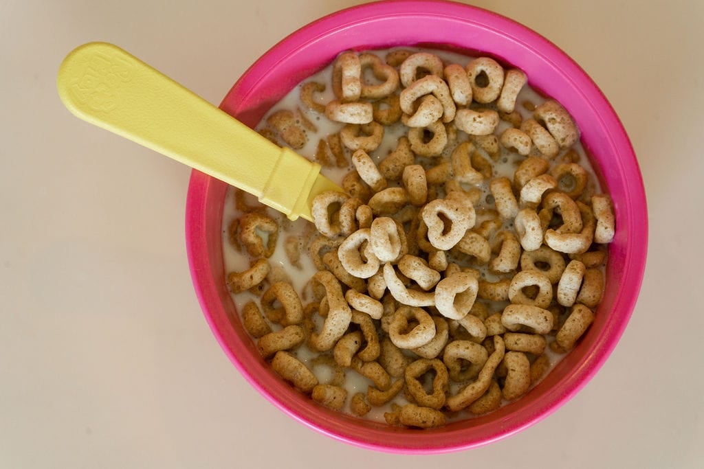 Pink kid's bowl filled with cheerios and milk with a yellow spoon sticking out.