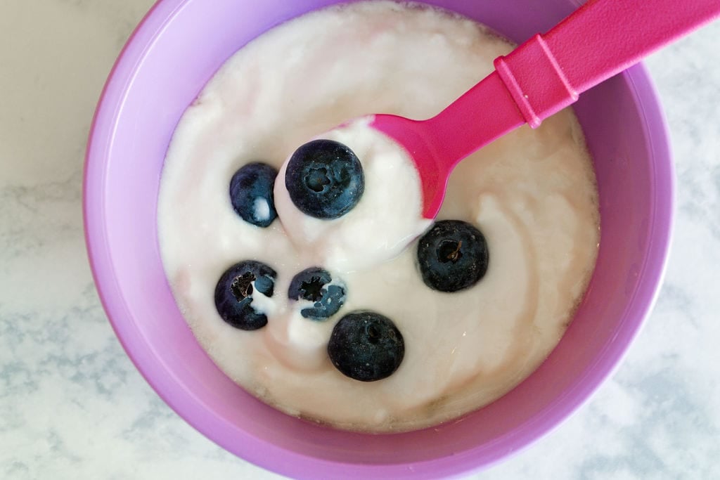 Purple bowl with yogurt and blueberries on top. Pink spoon scooping out some.