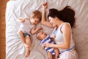 A woman lies on a bed smiling at a baby and a young child. The baby wears a striped onesie, and the child is in a tank top and shorts.
