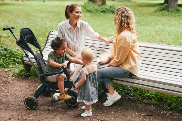 Full length portrait of two young mothers sitting on bench in park and playing with cute toddlers