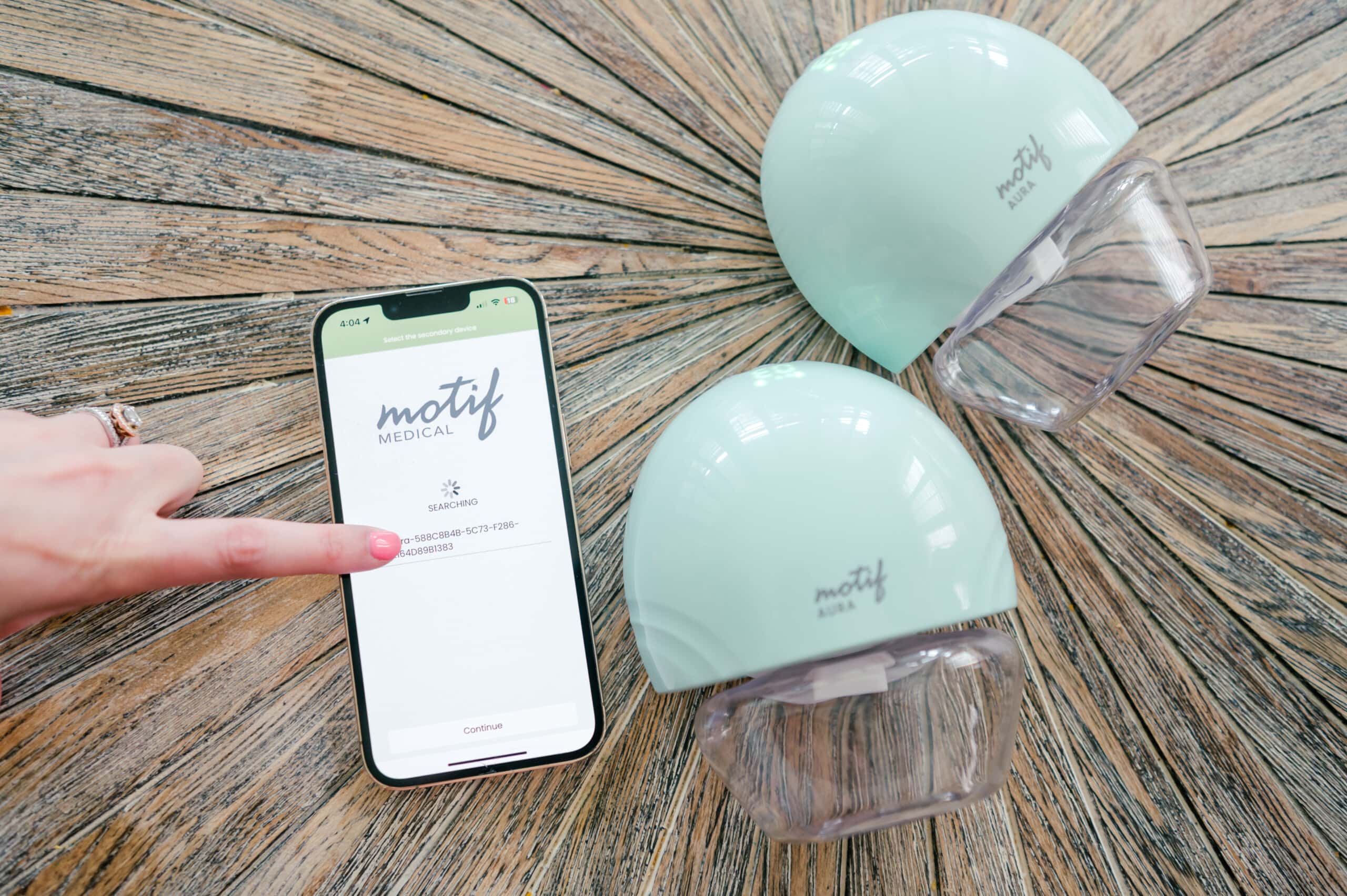 The Motif Aura breast pump on a wooden background with a smart phone showing the Motif app and a woman's hand pressing the app.