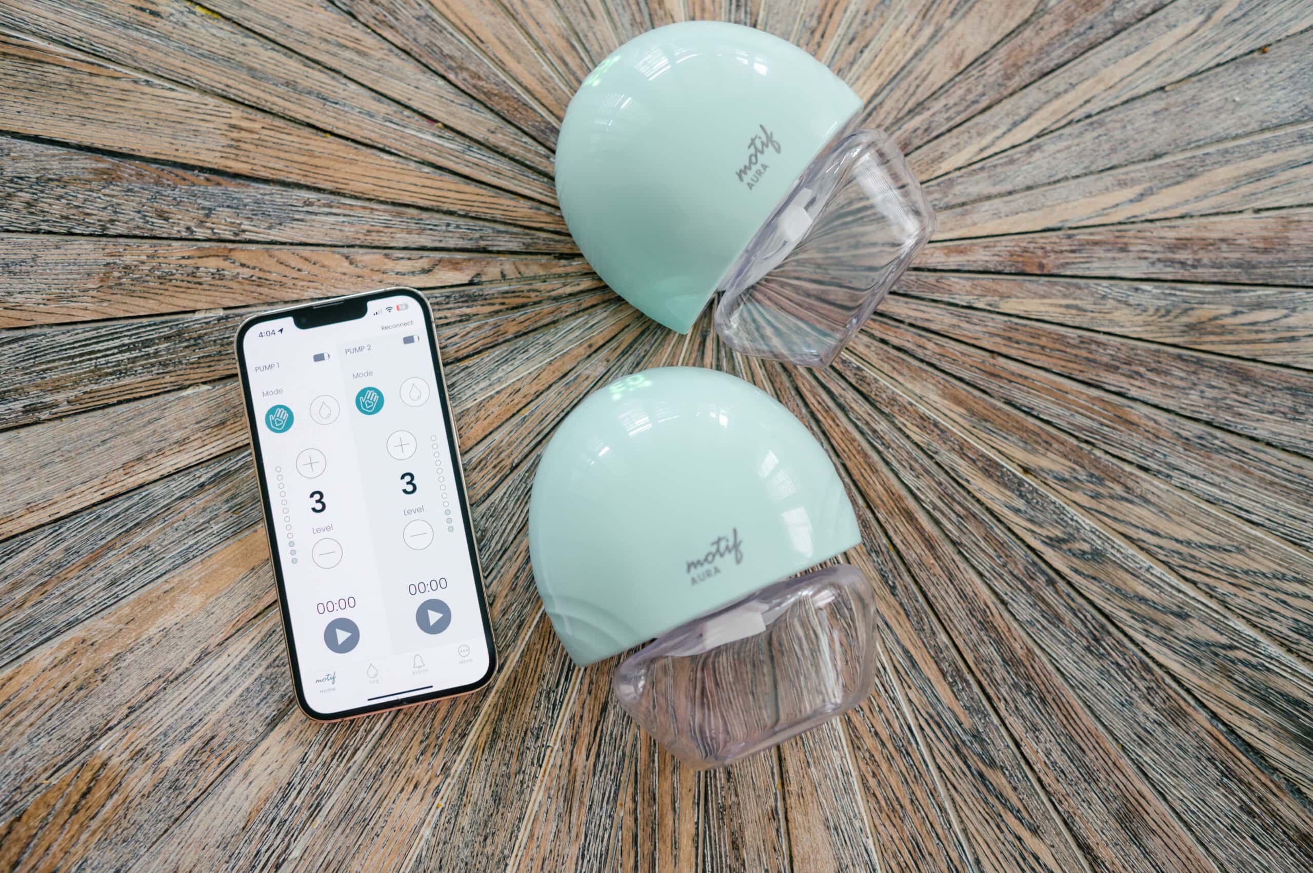 Motif Aura breast pumps on a wooden backdrop next to a smartphone with the Motif app on the screen