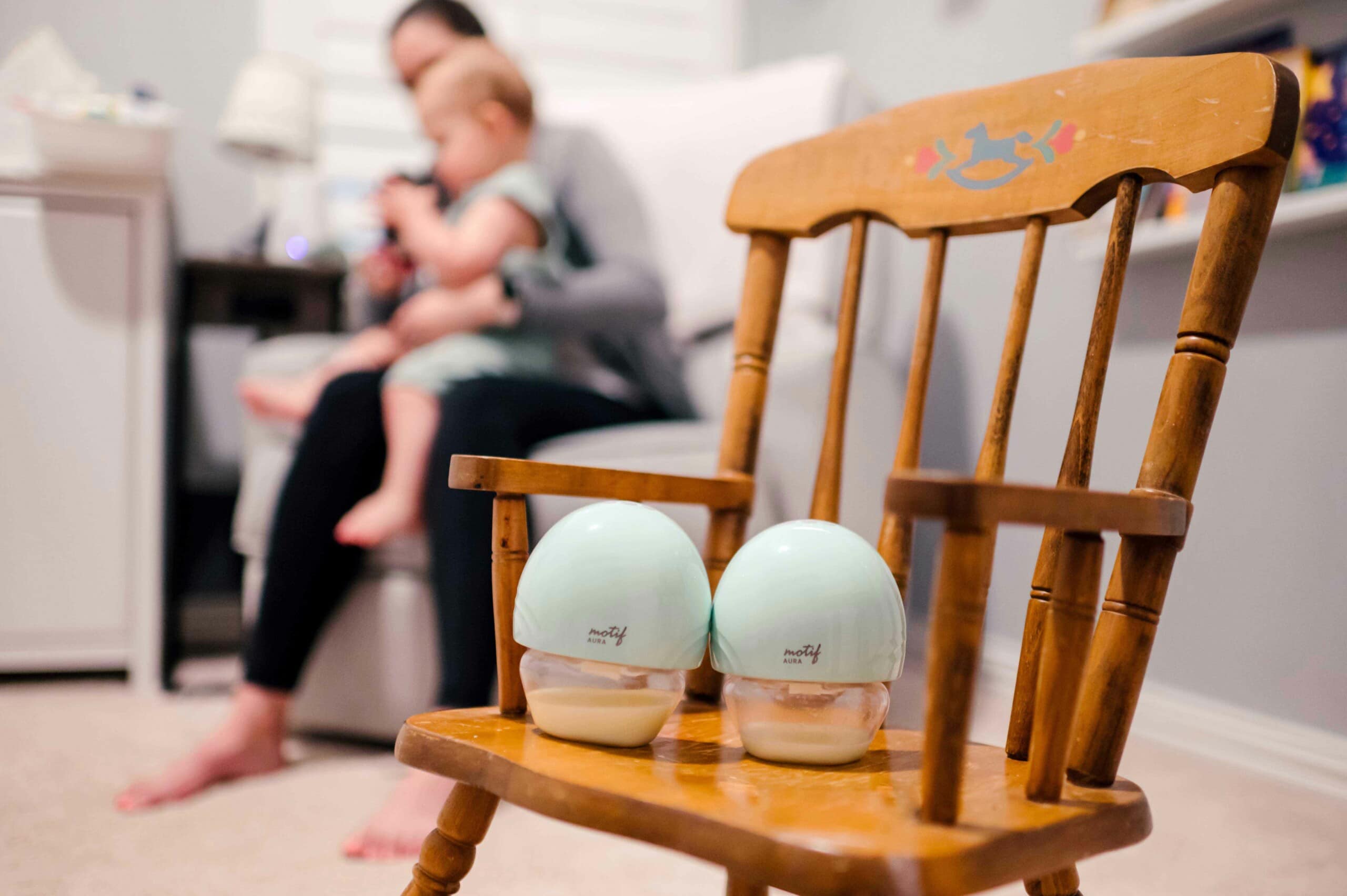mom and baby in the background. two Motif wearable breast pumps sitting on rocking chair.