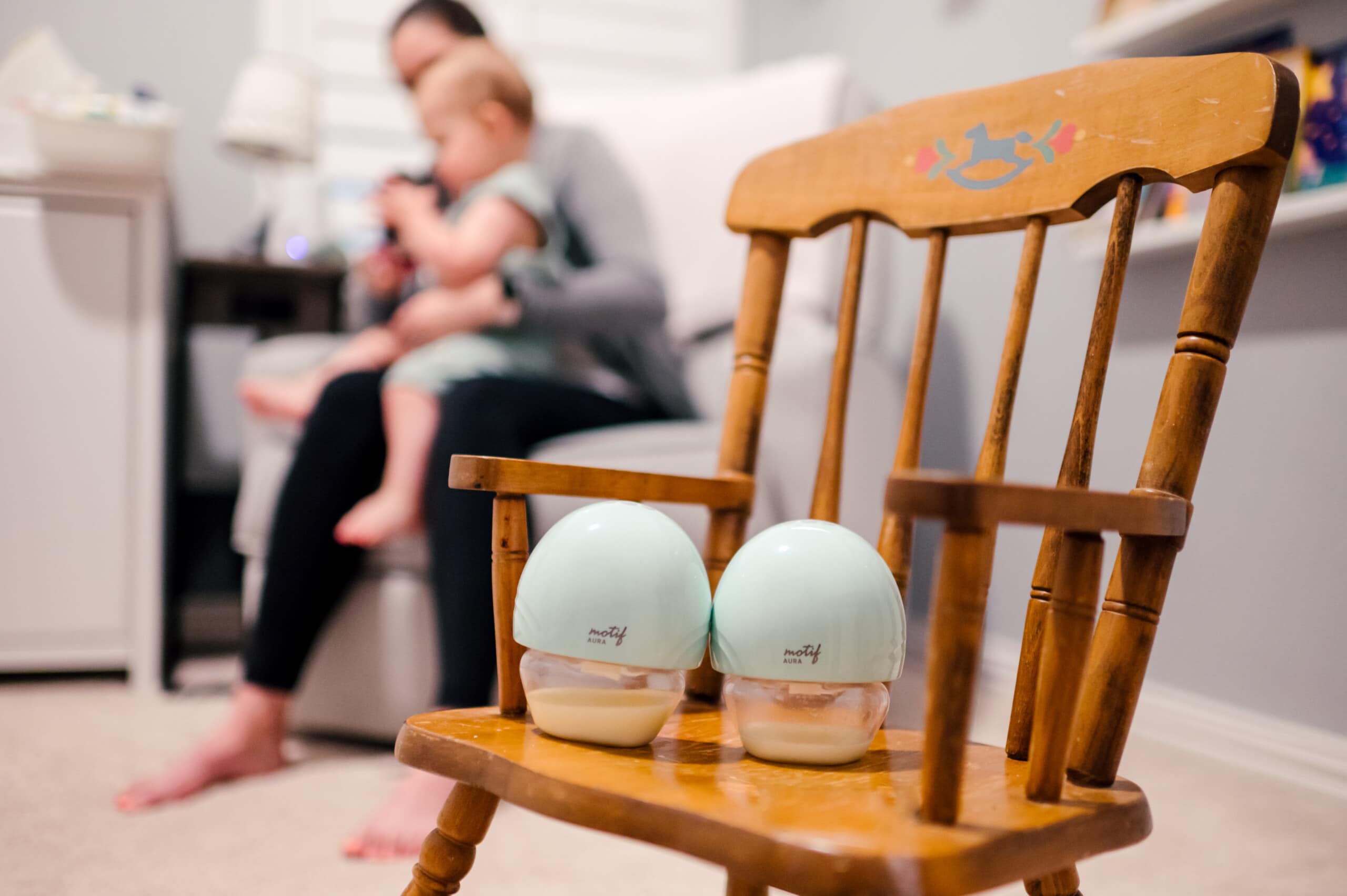 The Motif Aura breast pumps are sitting on a rocking chair in the foreground and a mom and her baby boy are sitting on the glider in the nursery in the background.