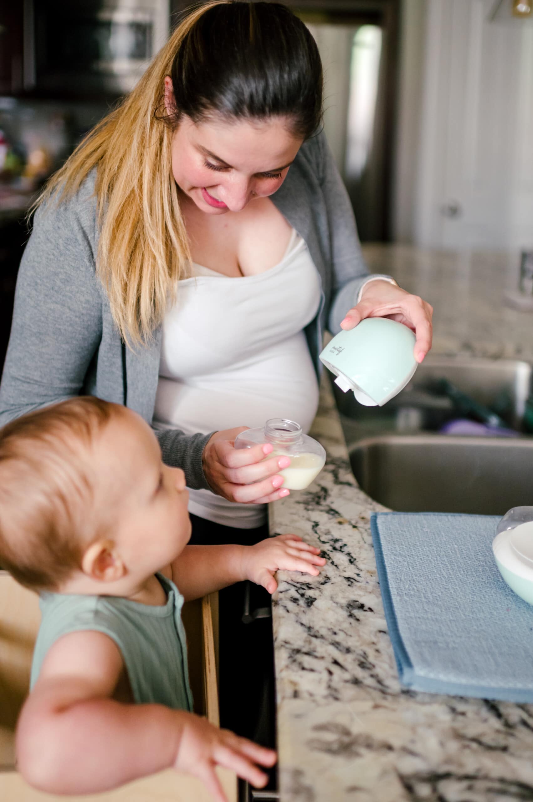Mom holding the Motif Aura breast pump with milk in it while she is standing in her kitchen with her toddler boy.