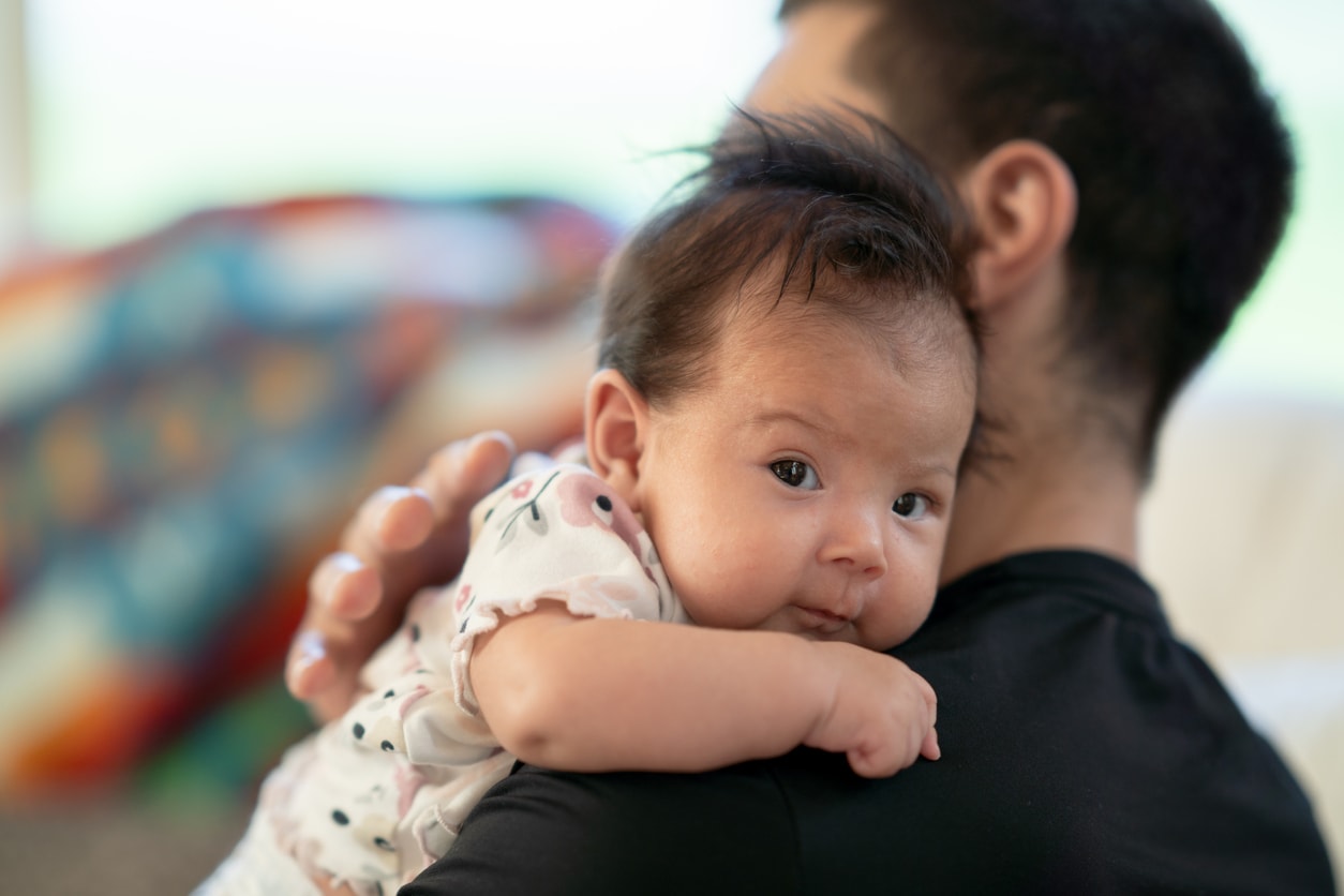 A sweet little newborn looks at the camera as she is propped up on her fathers shoulder while he is burping her. The sweet little indigenous baby has dark fluffy hair and is nestling into her fathers neck and shoulder.