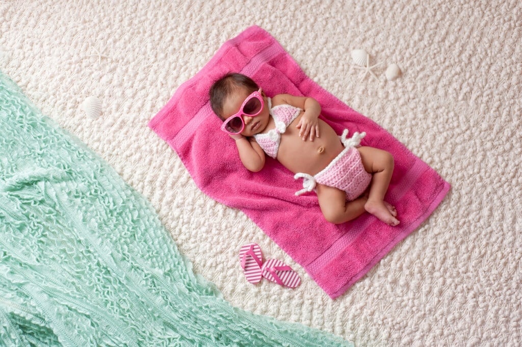 Four week old newborn baby girl sleeping on a pink towel. She is wearing a crocheted pink and white bikini and pink sunglasses. Shot in the studio with props made to look as if she's at a beach.