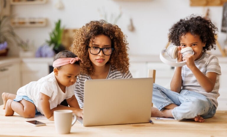 Young irritated stressed African-American mother covering ears and looking at laptop screen while working remotely from home with two small kids, being distracted from work by noisy naughty children