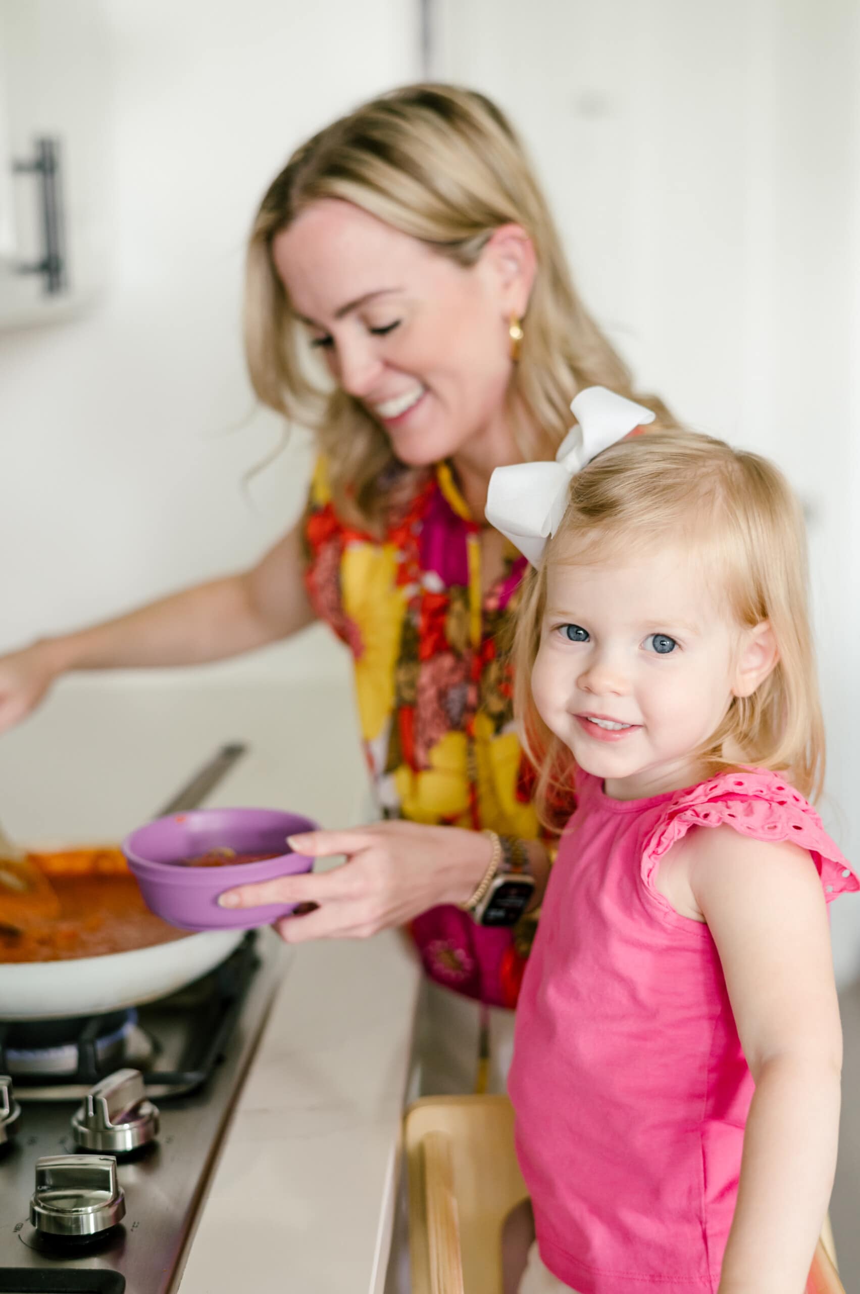 Young daughter smiling at the camera while her mom on the background is stirring pasta sauce at the stove.