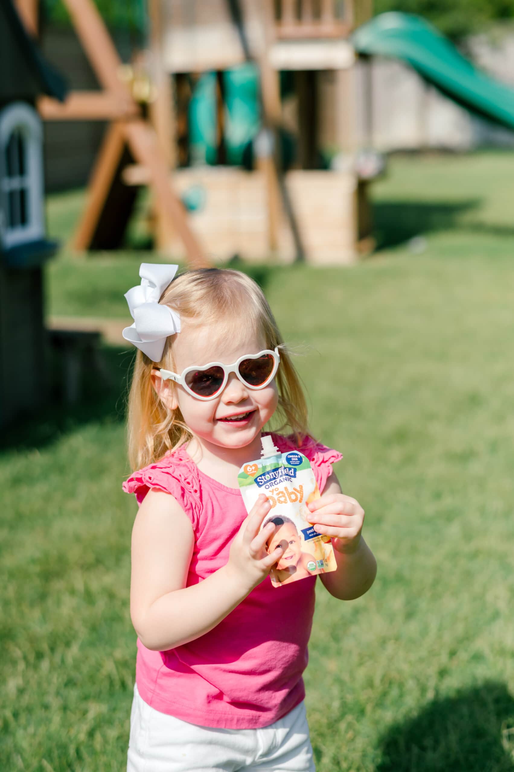 Little girl outside wearing heart-shaped sunglasses holding a Stonyfield Yobaby yogurt pouch.