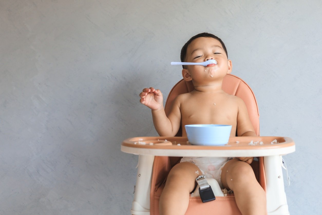 Happy infant Asian baby boy eating food by himself on baby high chair and making mess.