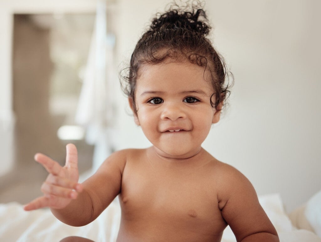 Baby, rock and roll hand sign, portrait and sitting in bed, morning and happy newborn in house. Child, innocent and play in bedroom, smile and happiness while relax, cute and sitting in room in home.