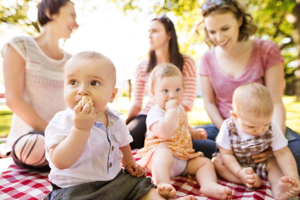 Close up of baby boy eating pie in public park having picnic, mothers with babies in the background.