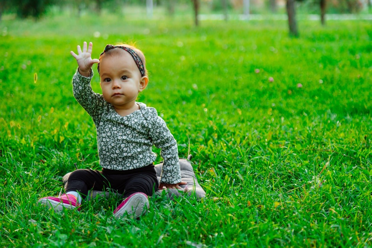 Happy baby waving hello sitting on green grass lawn
