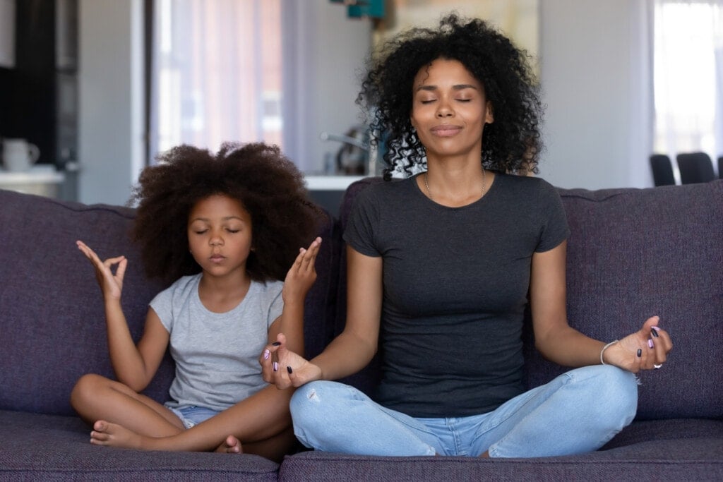 Mindful mom with cute funny kid daughter doing yoga exercise at home, sitting on the couch focusing on their breath and meditation. Mum teaching child to meditate.