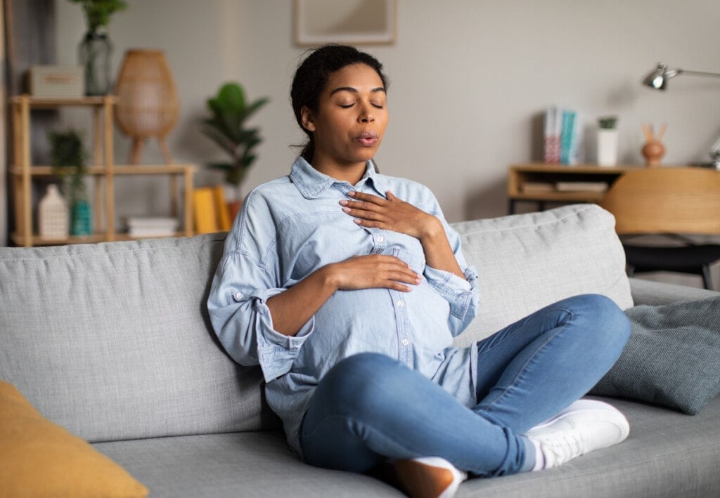 Pregnancy Yoga. Pregnant African American Woman Doing Breathing Exercise With Eyes Closed Relaxing Sitting On Couch At Home. Relaxation And Childbirth Healthcare Concept