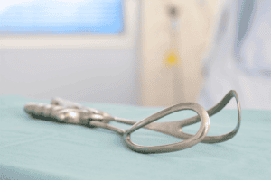 A close-up of a medical instrument, likely a vaginal speculum, resting on a light blue draped surface. The background appears to be a blurred hospital or clinical setting with blue and white tones, hinting at the preparation for procedures like forceps delivery.