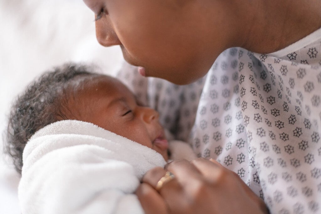 A young mother is kissing her newborn son on the forehead. They are at the hospital. The mother is wearing a hospital gown and the baby is wrapped tightly in a white blanket.
