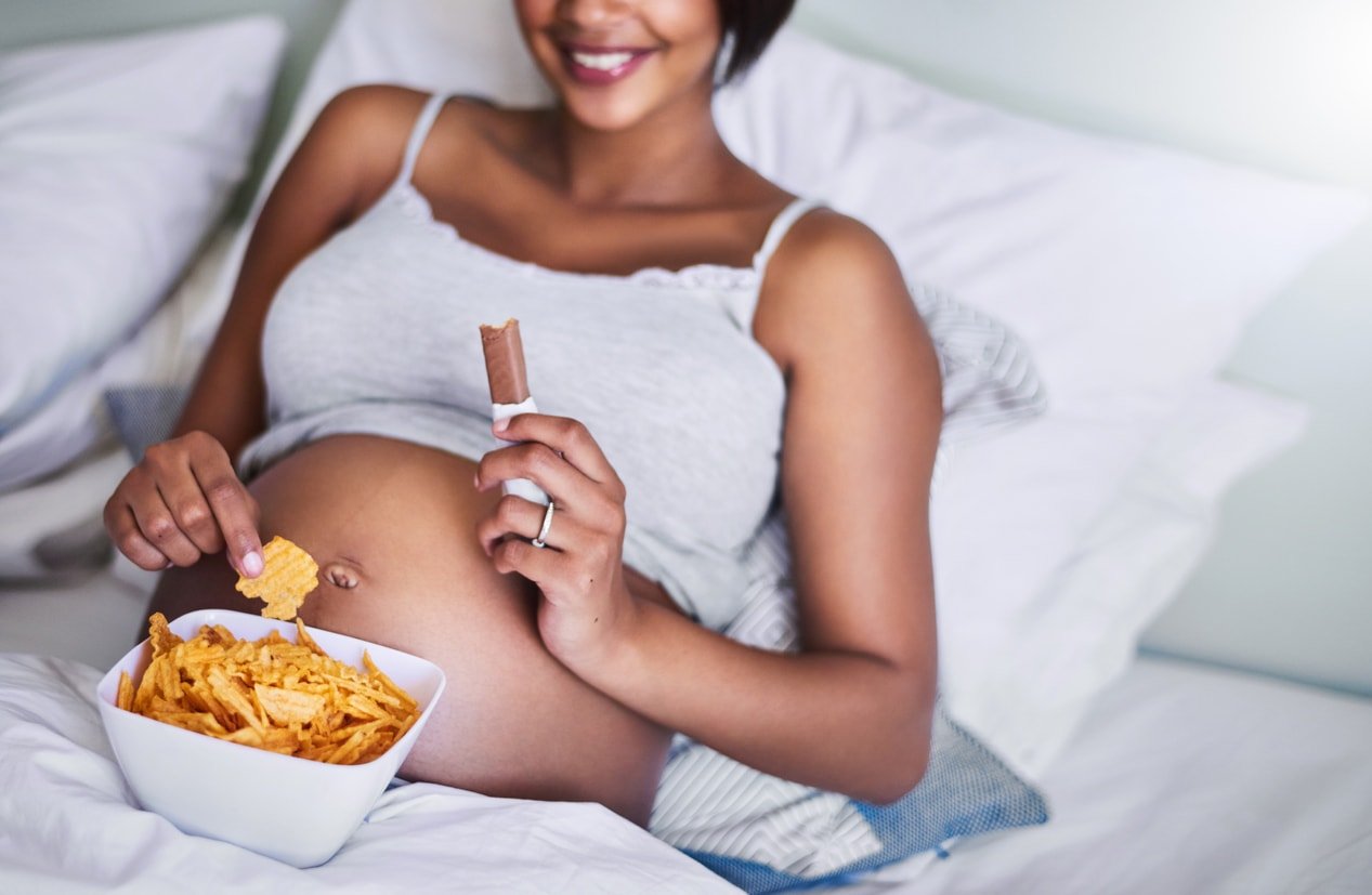 Cropped shot of a pregnant woman enjoying a chocolate and potato chips in bed