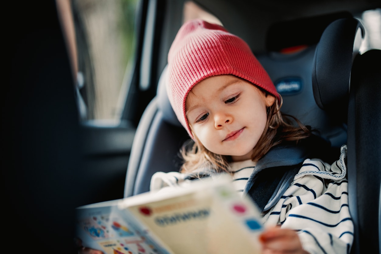 Smiling girl enjoy reading book while sitting in car safety seat.
