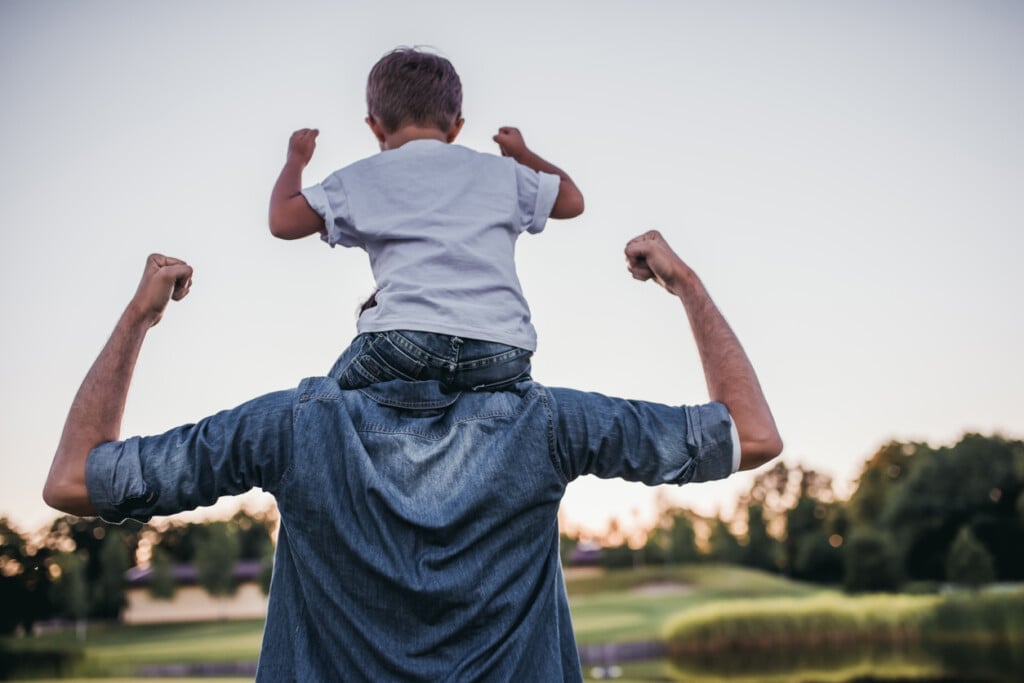 Dad and son having fun outdoors. Toddler son is sitting on his dads shoulders. It's a behind shot and they are both flexing their arms showing their muscles.