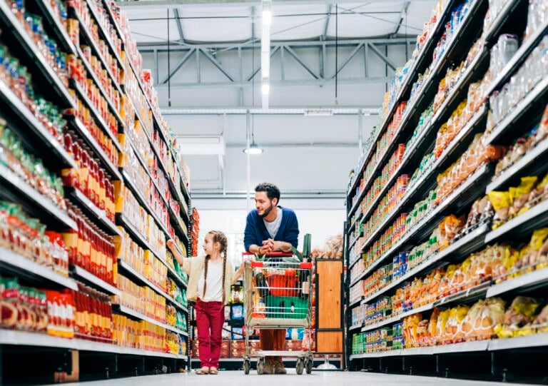 Girl buying groceries with father at supermarket. Man is leaning on shopping cart while daughter choosing food products. They are at grocery store.