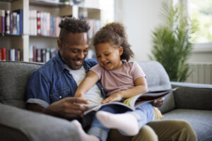 Happy African American father and adorable mixed race daughter are reading a book and smiling while spending time together at home. Children education and development concept