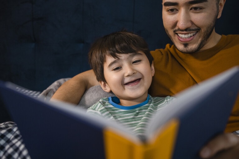 An Asian man and his young son wearing pyjamas in his bedroom on a winter's night. They are lying in bed while the father reads his son a bedtime story.