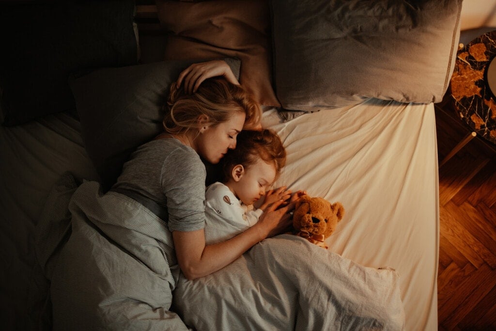 Beautiful mother and daughter sleeping together in bed. It is the evening and there is lamp on. Mom is holding her daughter and keeping her close. There is a teddy bear under the covers.
