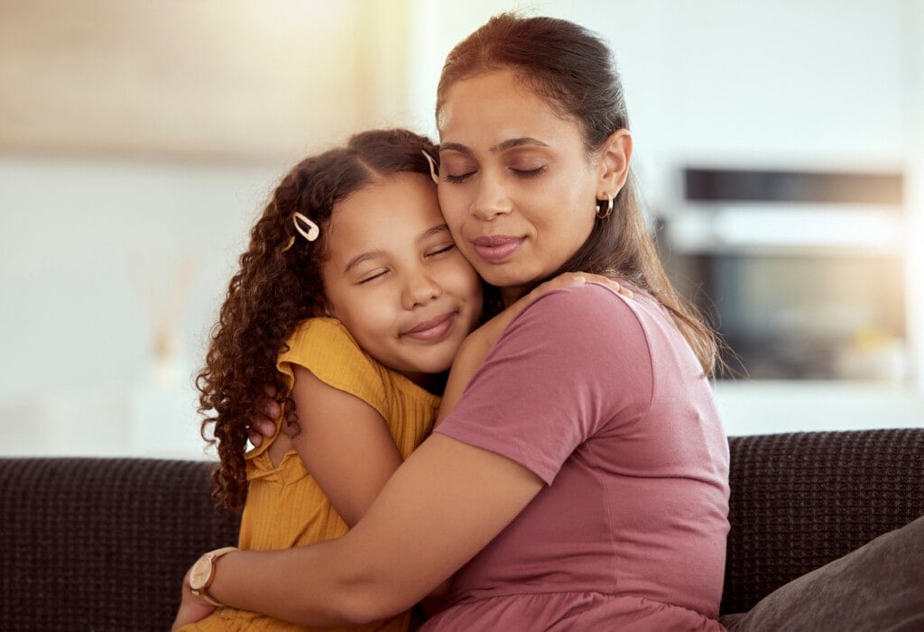 Mixed race single mother and daughter hugging in home living room. Smiling hispanic girl embracing and bonding with single parent in lounge. Happy affectionate woman and child together on weekend.