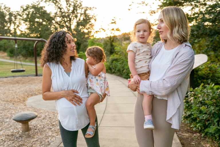 Two young mothers talk happily as they hold their toddler daughters on their hips and walk through the park at sunset. One of the women is eight months pregnant.