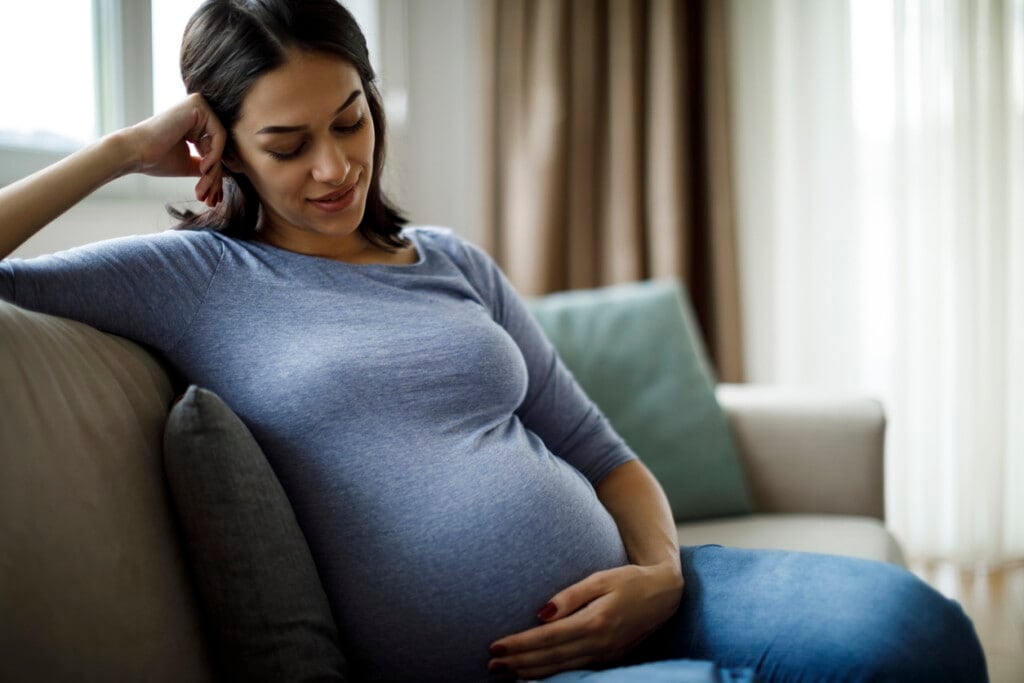 Portrait of young happy pregnant woman sitting on a couch looking down at her pregnant belly