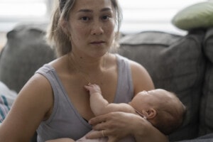 A middle aged mother sitting on her couch holds her baby in her arm with a tired and stressed expression.
