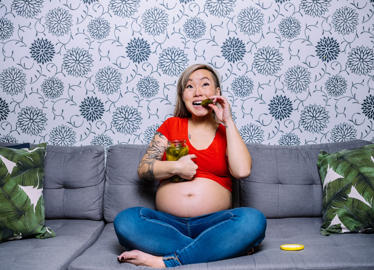 Young pregnant Asian woman sitting on the sofa against wall paper wall in living room. She is wearing jeans and red top with exposed pregnant belly. She is holding a jar of dill pickels and biting one. Her arms featuring tattoo art. Typical pregnancy cravings.
