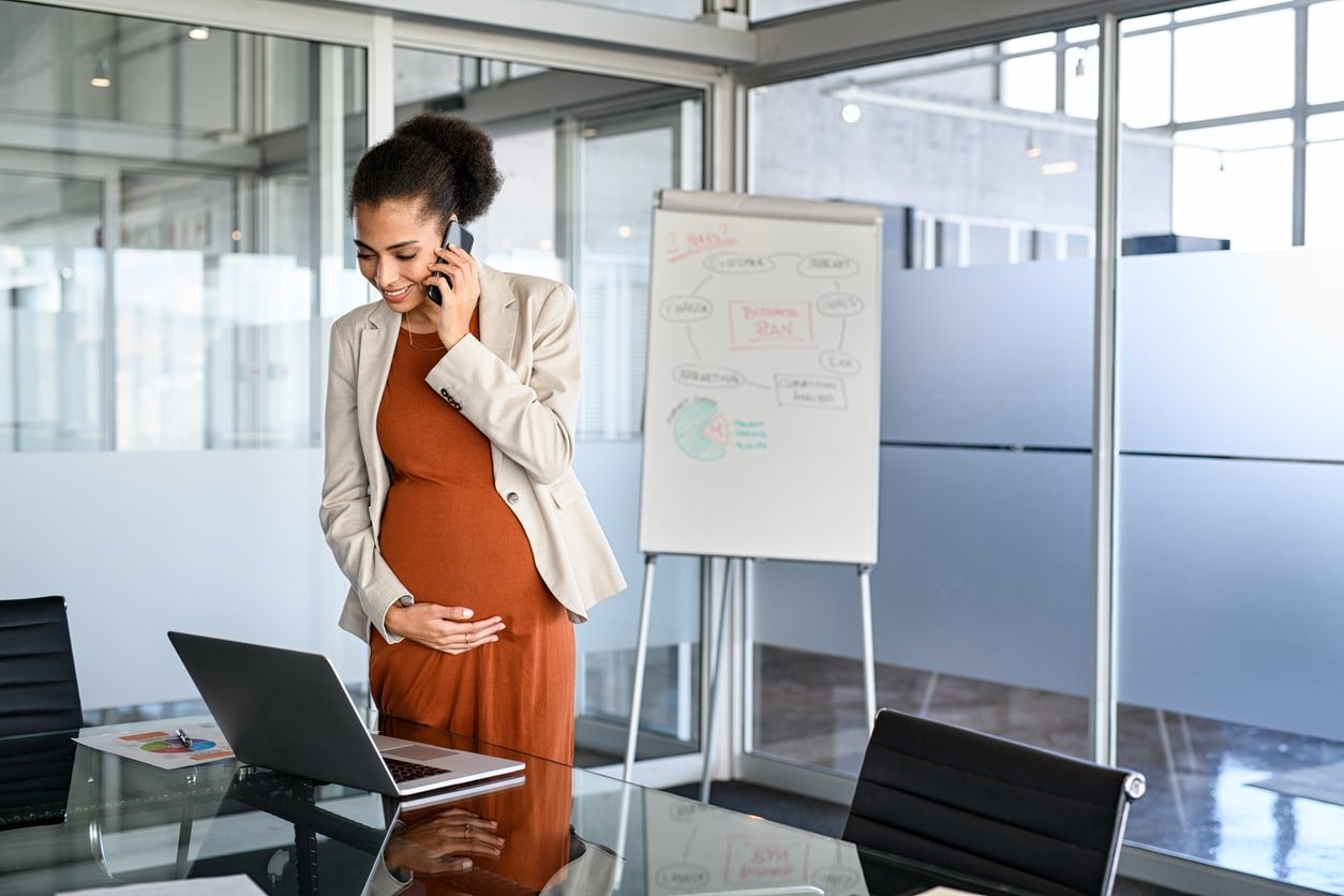 Smiling pregnant businesswoman touching belly while using laptop and talking over phone in modern office. Cheerful pregnant entrepreneur talking on phone while working with copy space. Beautiful african american woman working on laptop and talking on mobile phone.