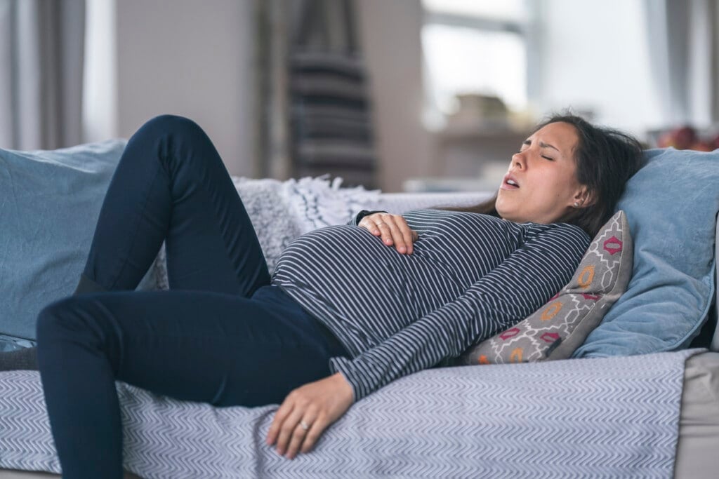 A mixed-race woman is pregnant. She is in her second trimester. The woman is lying on the couch in discomfort. She is breathing heavily and is resting her hand on her stomach. Prenatal, morning sickness, back pain, heartburn, and constipation concepts.