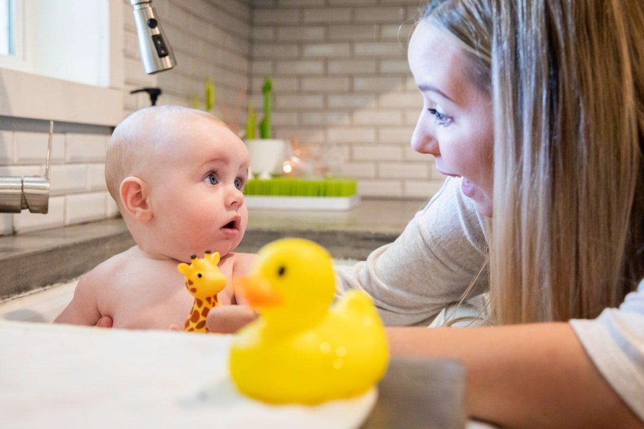 Young adult mother is giving her infant son a bath in the kitchen sink at home and talking to him while he plays with bath toys