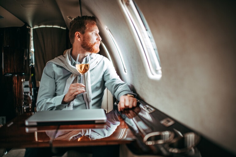 Young successful man drinking wine and enjoying himself while traveling with a private airplane. Laptop is on the table in front of him.