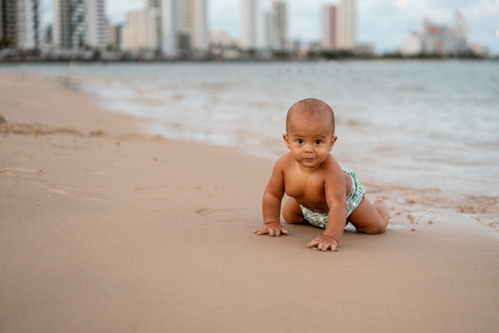 A baby crawling in the sand on the beach. The buildings are in the background.