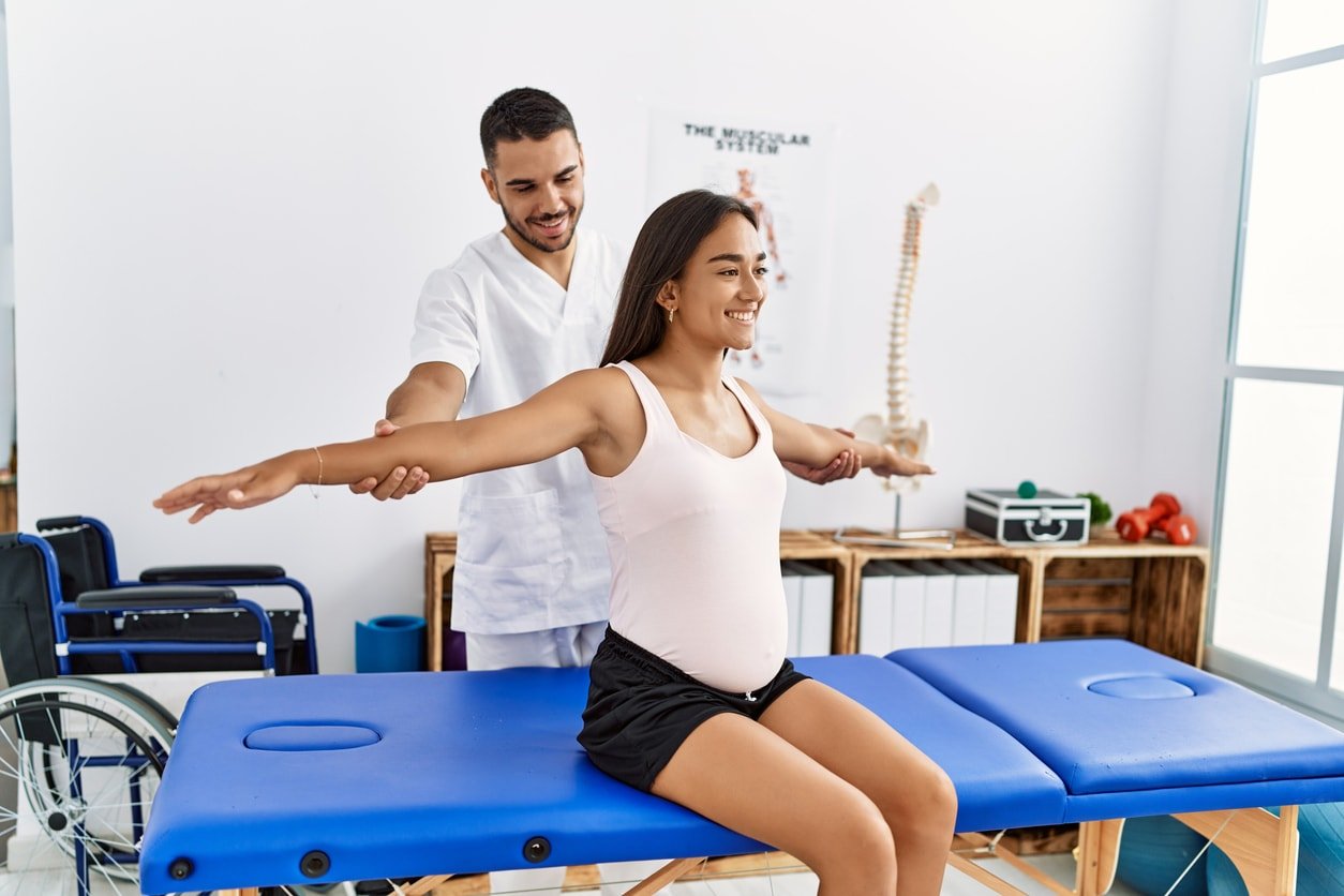 Latin man and woman wearing physiotherapist uniform having pregnancy rehab session at physiotherapy clinic
