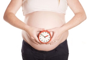 A pregnant person holds a small analog clock against their bare belly, indicating the anticipation of an overdue baby. The individual is wearing a light-colored top and dark pants, standing against a plain white background. The clock displays the time as approximately 12:04.