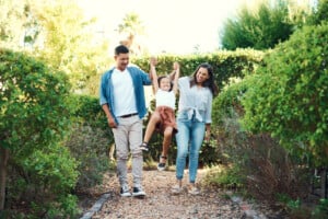 A man and a woman are walking in a garden, each holding one hand of a child between them. The child is lifted off the ground, smiling with joy as their parents embrace their responsibilities. The background is filled with greenery and sunlight filters through the trees.