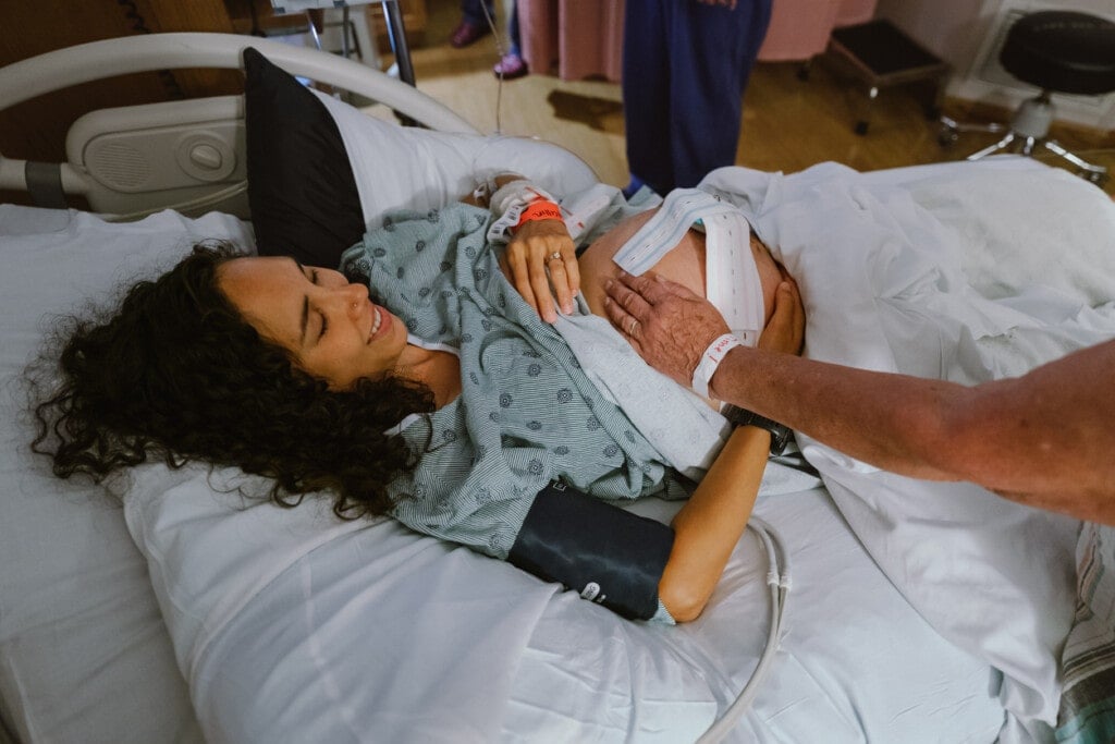 A pretty Eurasian woman who is in labor lies in a hospital bed and smiles as her mother affectionately rests her hand on her pregnant belly.