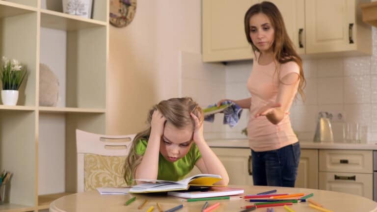 A young girl with long blonde hair sits at a table with her head in her hands, looking frustrated over an open book. Colored pencils are scattered on the table. A helicopter mom stands behind her, gesturing with one hand while holding a dish towel in the other.