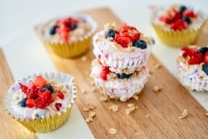 Six yogurt parfait cups with granola, blueberries, and strawberries sit on a wooden board, each in a gold or white paper liner.