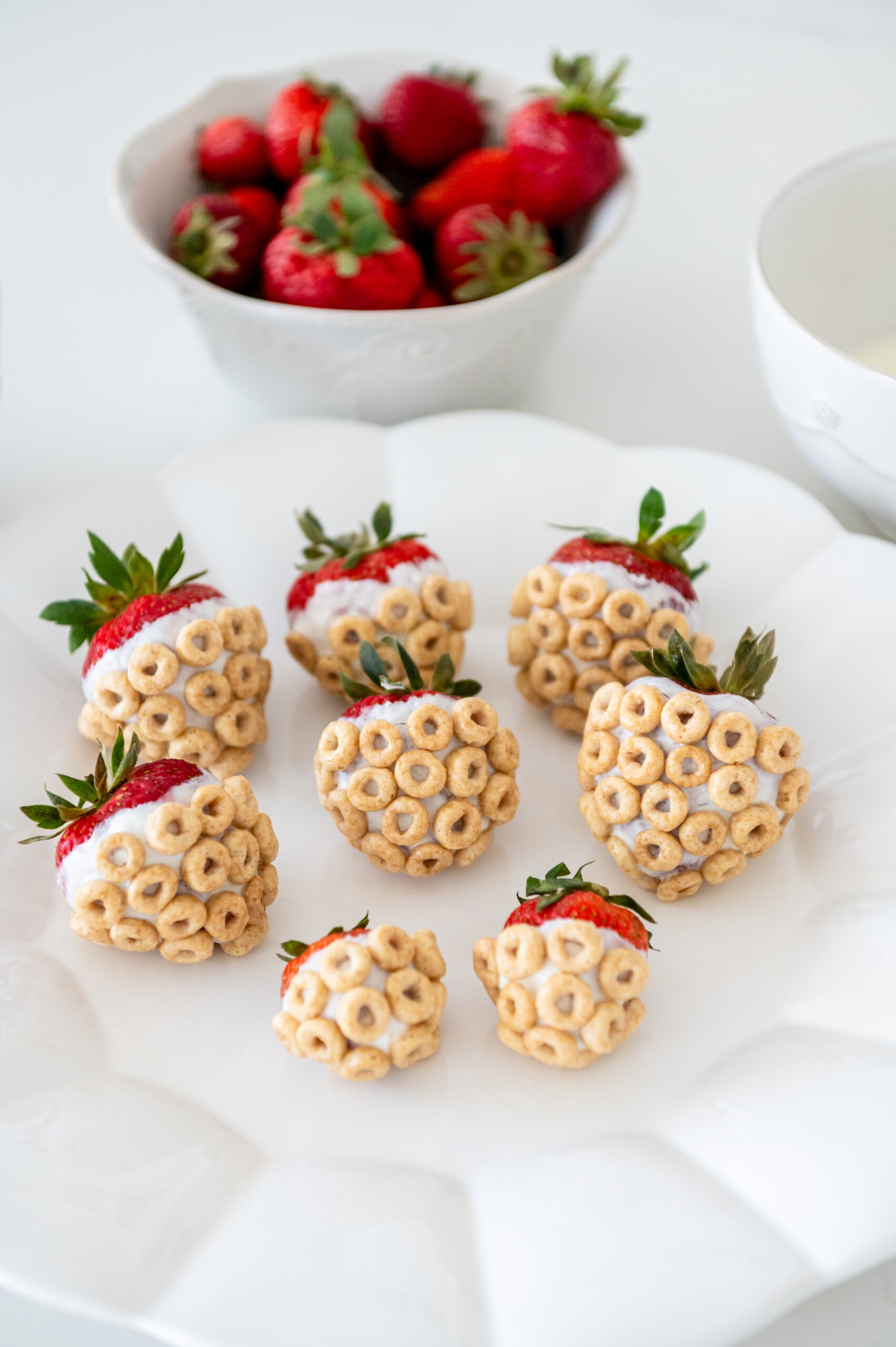 A plate with cheerio and yogurt-covered strawberries. There is a bowl of strawberries in the background.