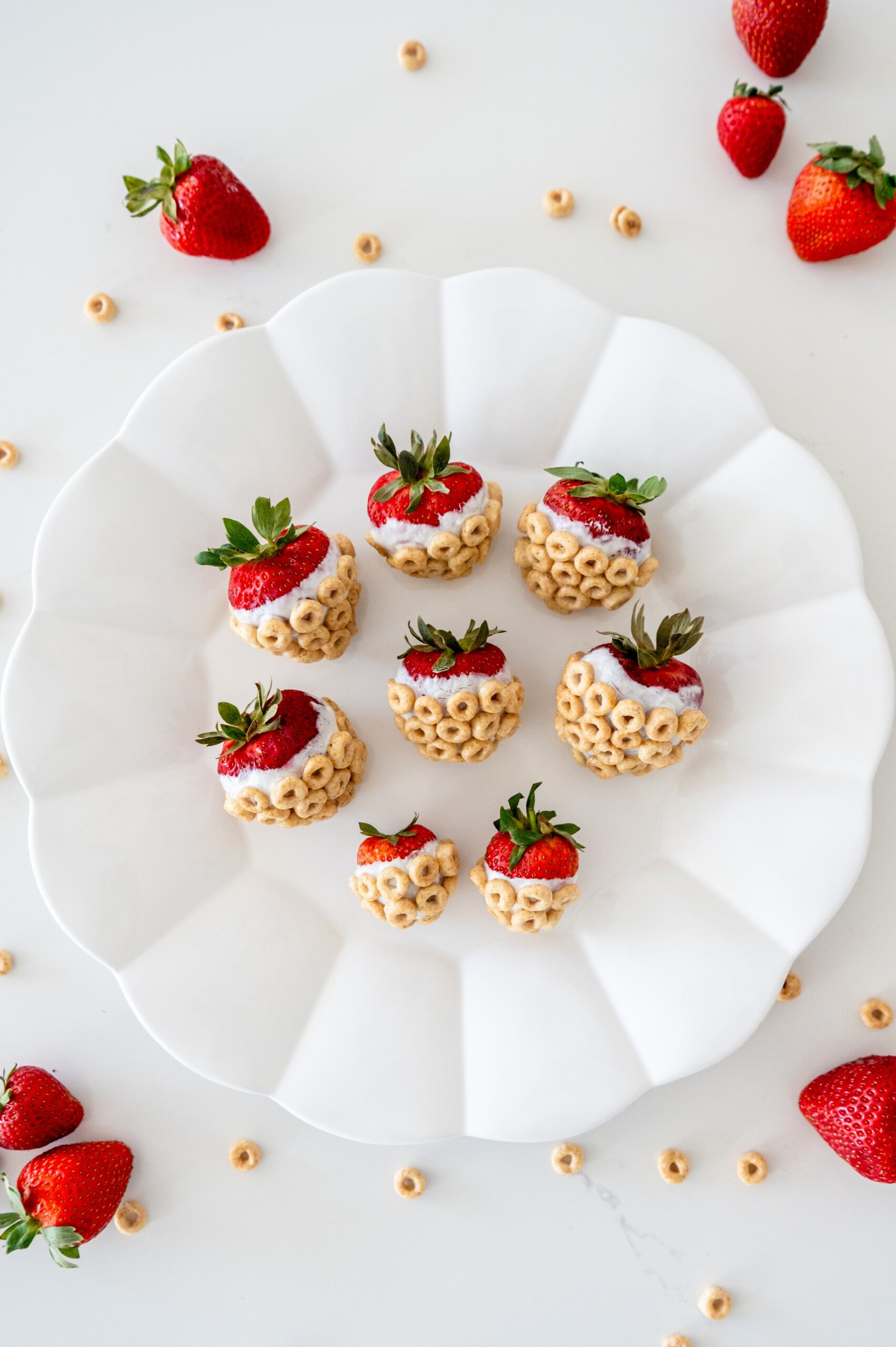 Strawberries dipped in yogurt and covered with Cheerios on a white plate in a white kitchen. There are strawberries and Cheerios scattered around.